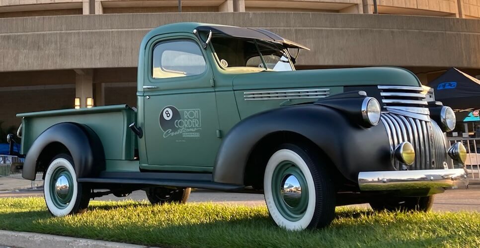 Ron Corder Customs An old green pickup truck is parked in front of a stadium.